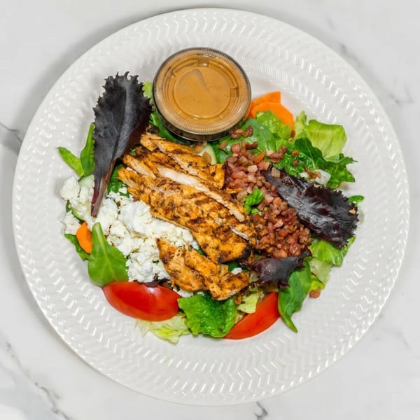 Grilled chicken salad with mixed greens, tomatoes, feta cheese, bacon bits, and a side of dressing.
