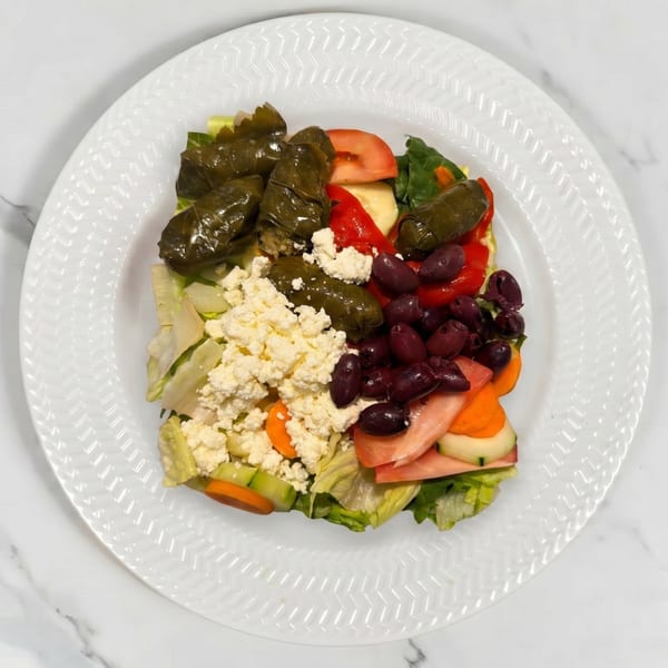 A vibrant Greek salad with stuffed grape leaves, feta, olives, tomatoes, and cucumbers on a white plate.